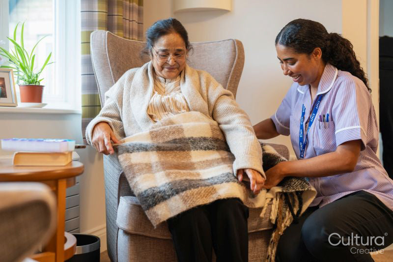 Nurse covering senior lady with blanket while she sits in armchair at nursing home