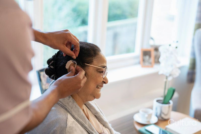 Nurse helps senior lady putting on hearing aid at nursing home