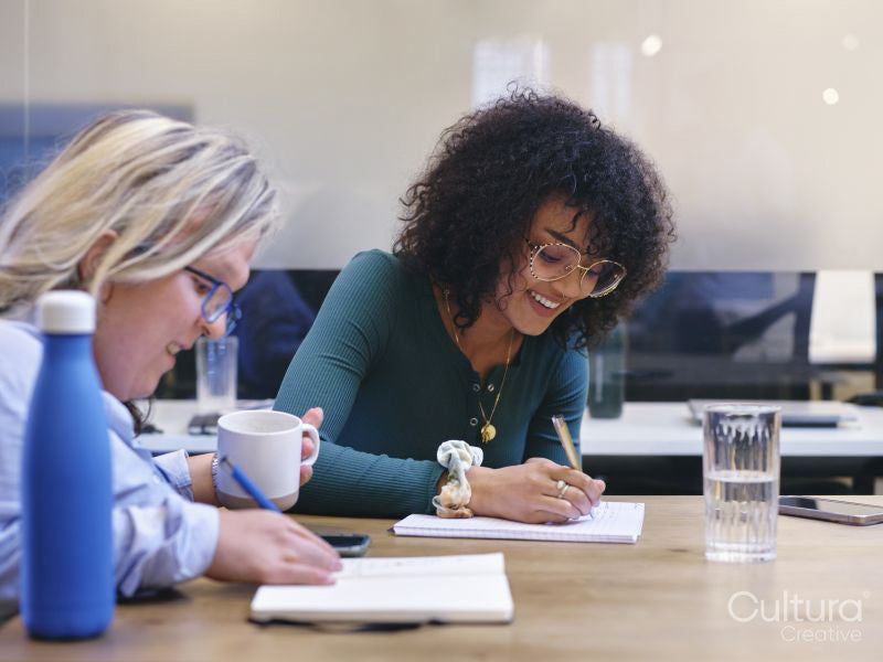 Two smiling women writing in notebooks in modern office