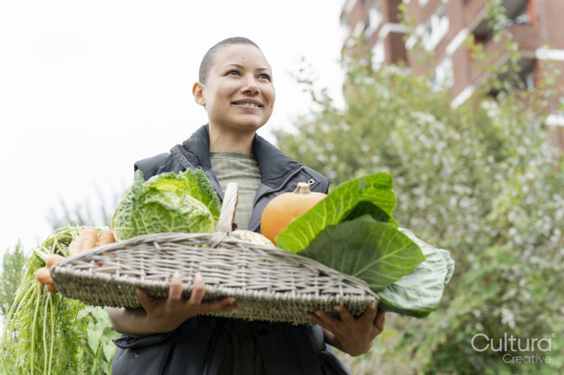 Portrait of smiling woman holding basket with fresh vegetables in urban garden