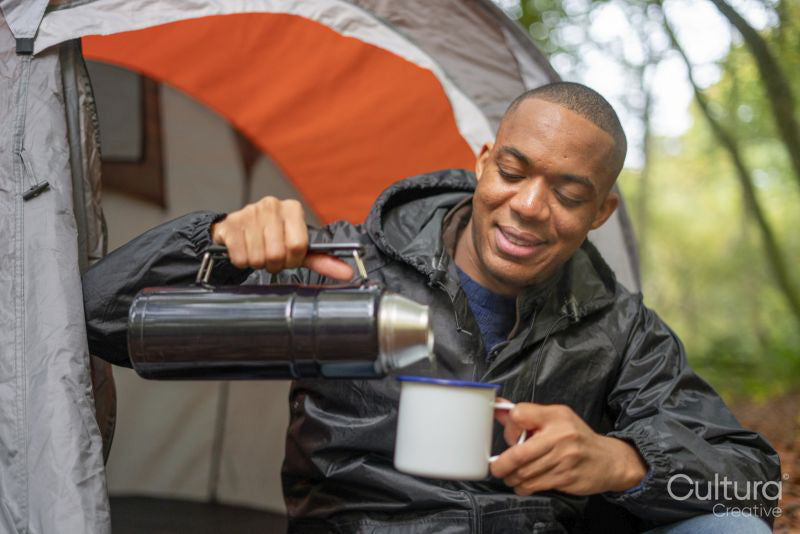 Man pouring warm drink into metal mug outside of tent