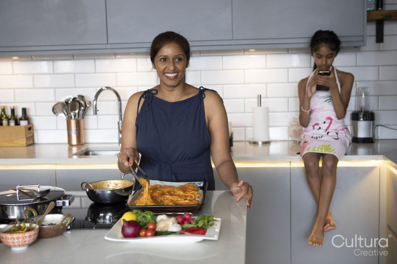 Smiling woman cooking in kitchen, daughter looking at smart phone