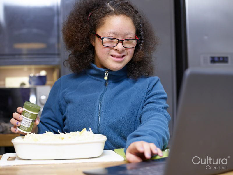 Girl with Down Syndrome using laptop and preparing food in kitchen