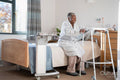 Smiling senior woman in bathrobe sitting on bed and holding mobility walker in nursing home