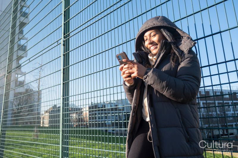 Portrait of smiling woman in overcoat with hood looking at smart phone at fence