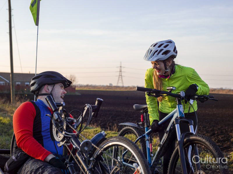 Disabled man on handcycle and woman with bicycle laughing in rural landscape