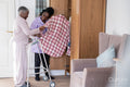 Smiling caregiver assisting senior woman standing at mobility walker and choosing shirt at closet