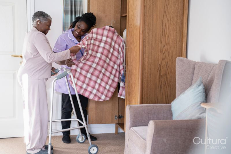 Smiling caregiver assisting senior woman standing at mobility walker and choosing shirt at closet