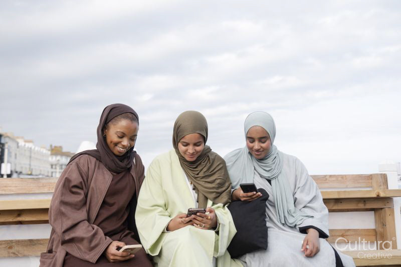 Three Muslin women using smart phones while sitting on bench