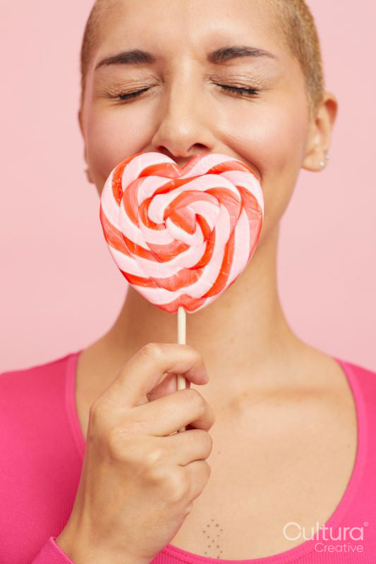 Young blond woman shielding mouth with heart shape lollipop, eyes closed