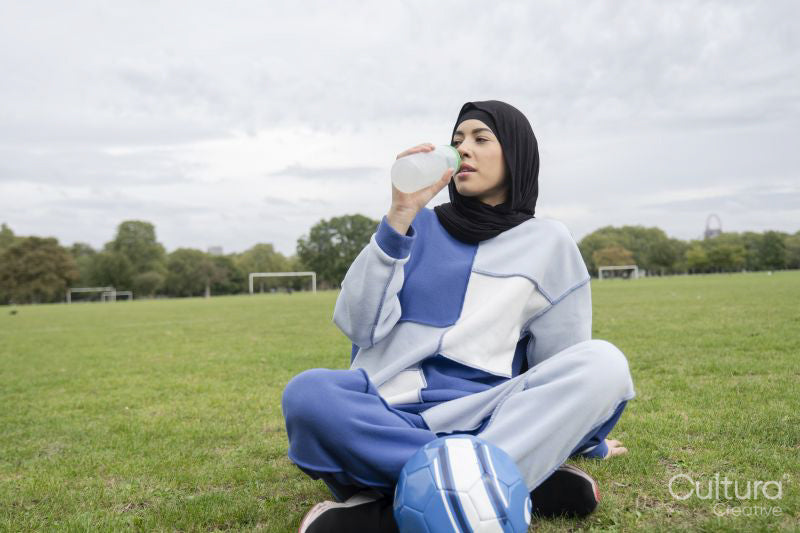 Portrait of woman in hijab sitting in soccer field, drinking water from bottle
