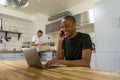 Man using laptop and phone in kitchen, boyfriend in background