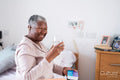Smiling senior woman in pajamas sitting on bed, holding pill organizer and glass of water