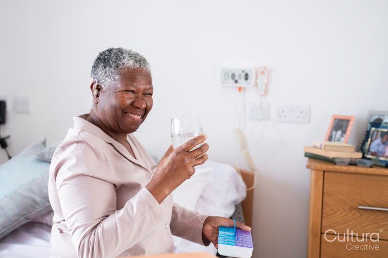 Smiling senior woman in pajamas sitting on bed, holding pill organizer and glass of water