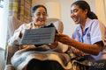 Senior lady sitting in armchair and showing tablet to nurse at nursing home