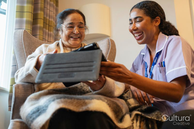 Senior lady sitting in armchair and showing tablet to nurse at nursing home