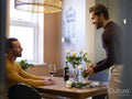 man arranging rose flowers in vase while boyfriend drinking wine