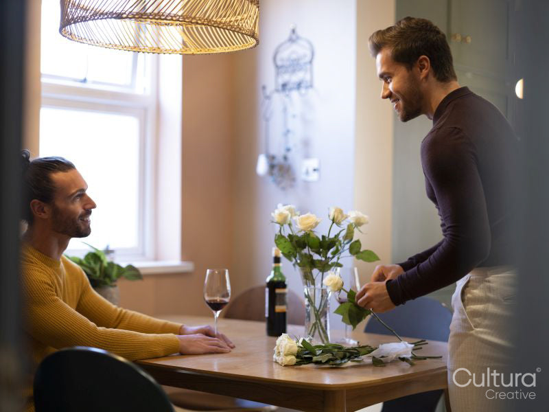 man arranging rose flowers in vase while boyfriend drinking wine