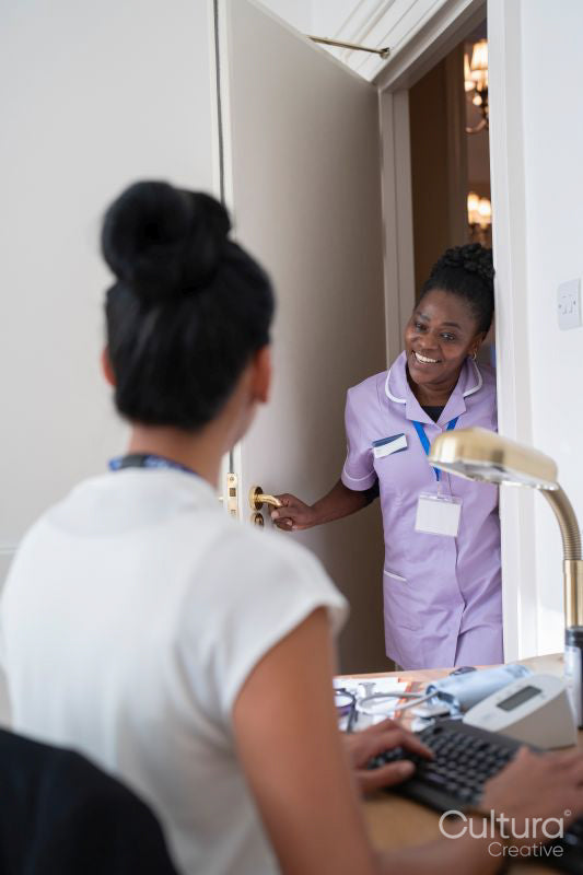Smiling nurse standing at door and talking to female doctor sitting at desk in office