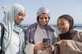 Three Muslim women looking at smart phone while standing on pier on sunny day