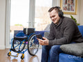 Man with headphones and smart phone sitting on sofa, wheelchair in background
