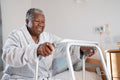 Smiling senior woman in bathrobe sitting on bed and holding mobility walker in nursing home