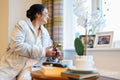 Senior lady sitting with phone and looking through window in bedroom at nursing home