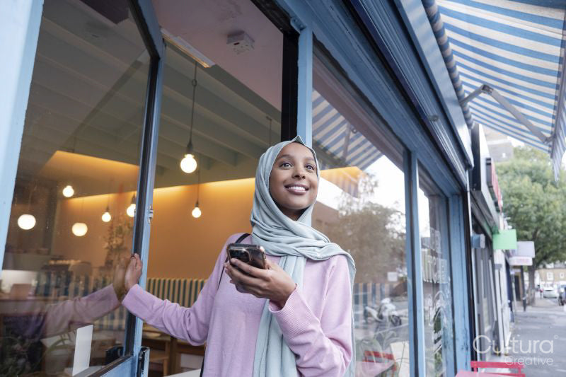 Young woman in hijab using phone while entering cafe