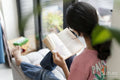 Girl sitting at hassock near window and reading book
