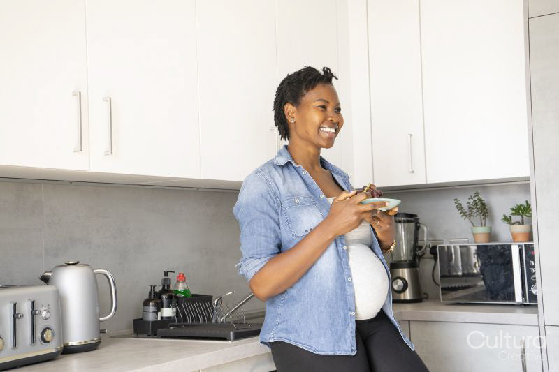 Smiling pregnant woman holding plate with red grapes in kitchen
