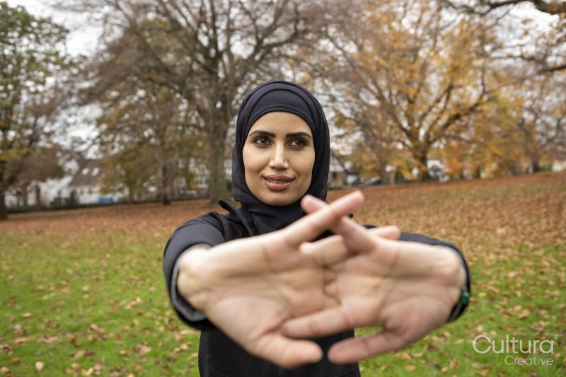 Smiling woman in black sports clothing and hijab stretching in park