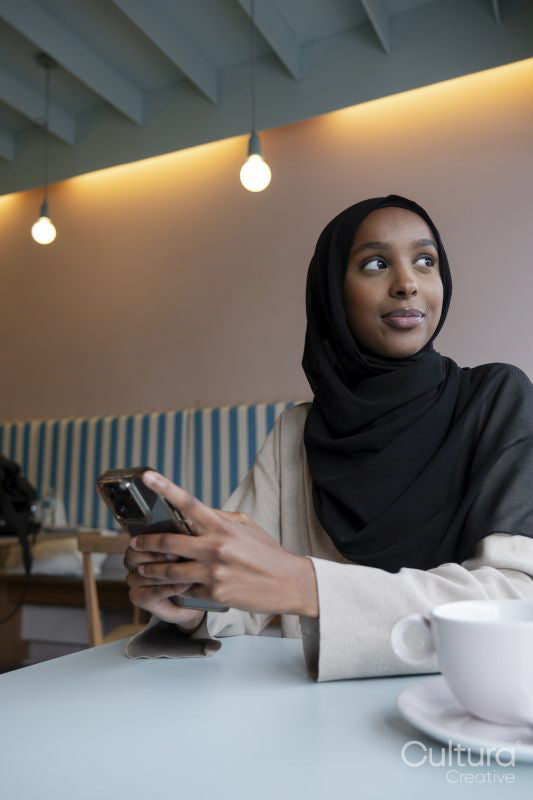 Smiling young woman in hijab using phone in cafe