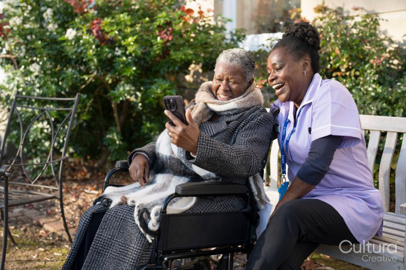 Smiling nurse and senior woman in wheelchair relaxing in park and looking at phone