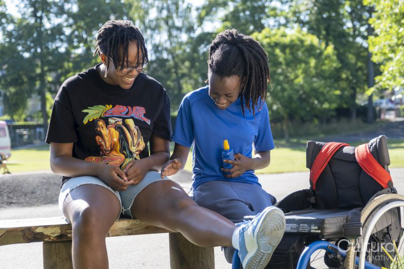Teenage girl in wheelchair with friend applying sunscreen in park