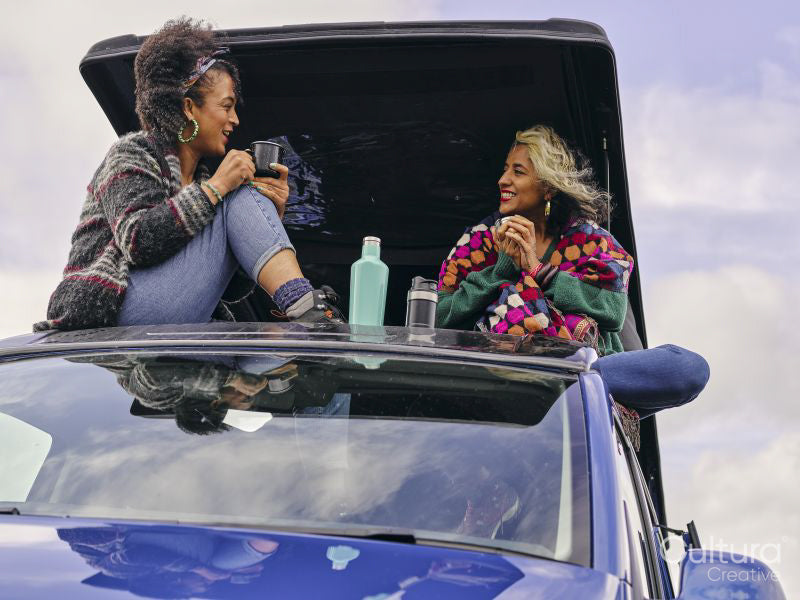 Women sitting on car roof and drinking from insulated drink containers