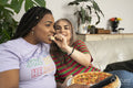Two young women watching tv and eating pizza