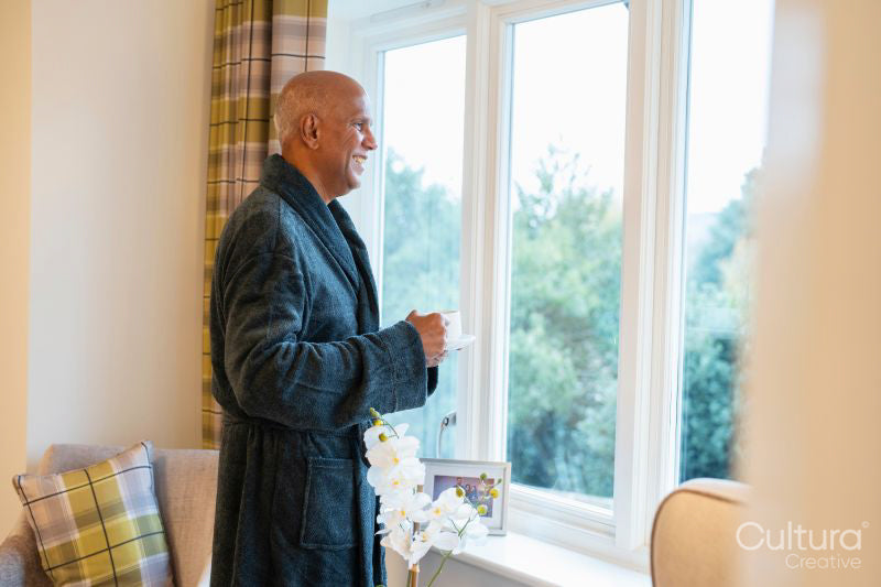 Senior man standing with coffee and looking through window in his room at nursing home