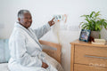 Smiling senior woman in bathrobe sitting on bed and pushing emergency button on wall