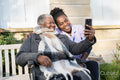 Smiling nurse and senior woman in wheelchair relaxing in park and looking at phone