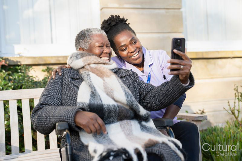 Smiling nurse and senior woman in wheelchair relaxing in park and looking at phone
