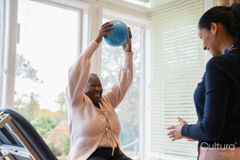 Female therapist assisting senior woman exercising with ball