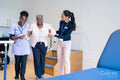 Female therapist and nurse supporting senior woman walking in exercise room