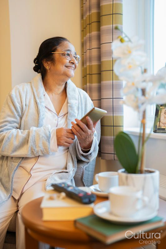 Senior lady sitting with phone and looking through window in bedroom at nursing home