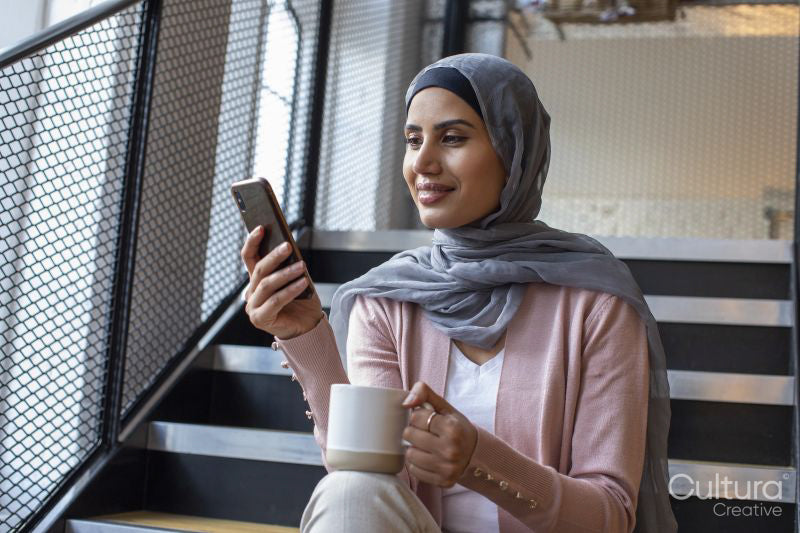 Smiling woman wearing headscarf sitting on steps and holding cup and smart phone