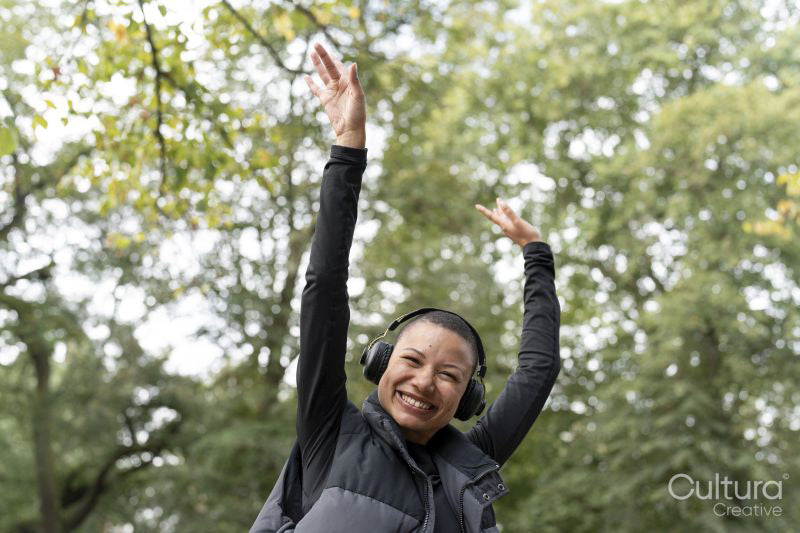 Portrait of smiling woman with headphones raising arms in park