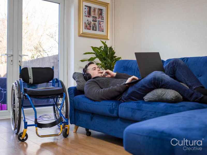Man with headphones and laptop lying on sofa next to wheelchair