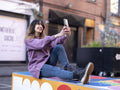 Woman taking selfie while sitting on wall in city street
