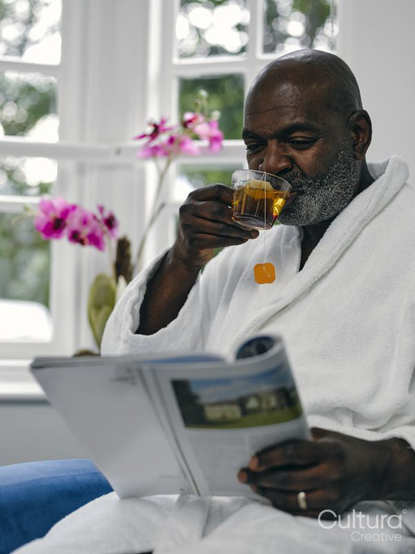 Mature man in white bathrobe reading magazine and drinking tea