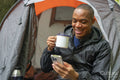 Smiling man using smart phone outside of tent and holding metal mug