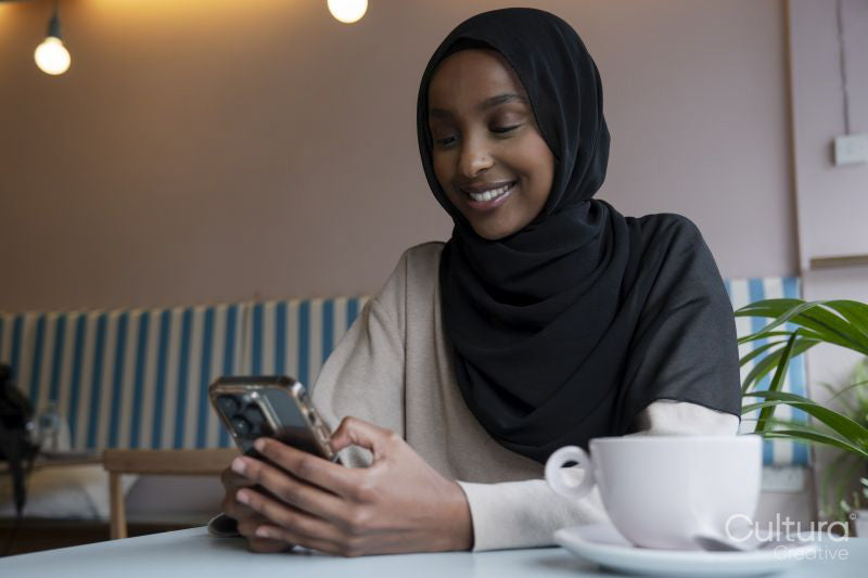 Smiling young woman in hijab using phone in cafe